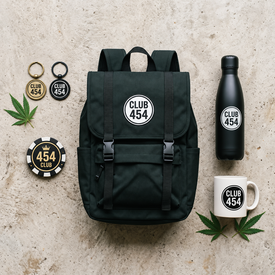Top-down flat lay of Club 454 merch on light concrete: two keychains, a brass door knocker, a black water bottle, a white mug and a black backpack