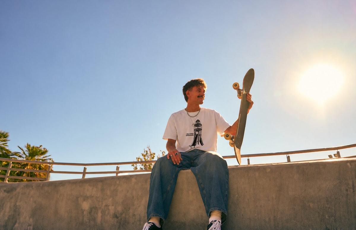 Young man holding a skateboard up against a bright sky, wearing a graphic tee